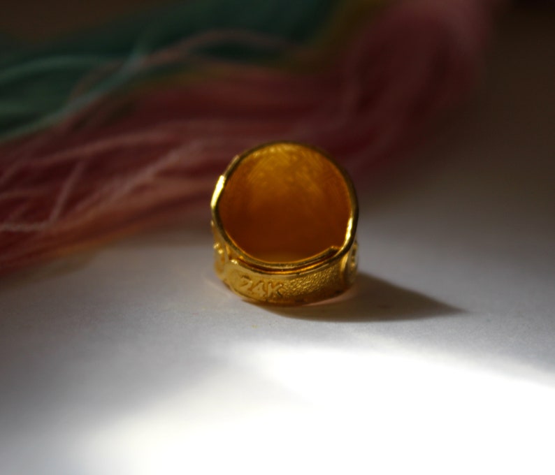Puede incluir: Primer plano de un anillo de oro con superficie texturizada. El anillo est&aacute; inscrito con "14K" y presenta un dise&ntilde;o detallado alrededor de la banda. El anillo se encuentra sobre un fondo suave y borroso con tonos rosas y verdes.