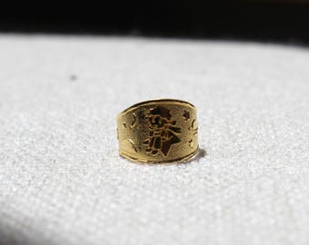 Puede incluir: Un anillo dorado con un grabado detallado de una figura, estrellas y lunas crecientes. El anillo está sobre un fondo blanco texturizado. El diseño del anillo es visible y el metal tiene un acabado brillante.