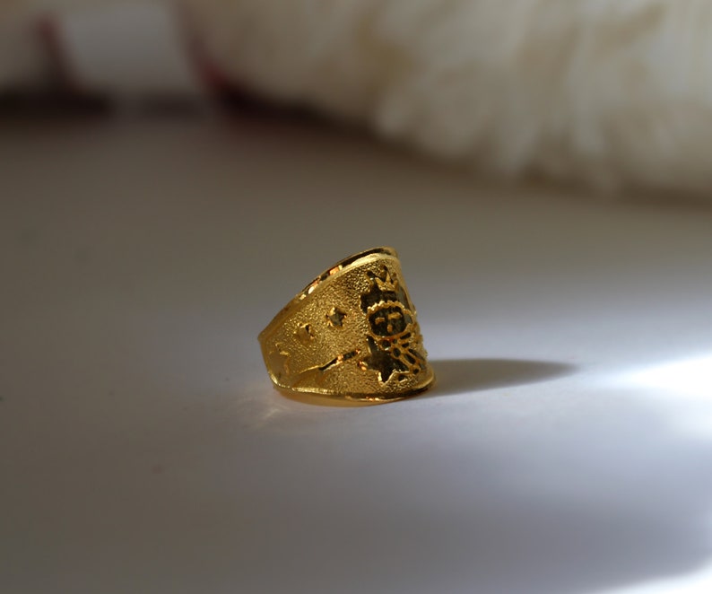 Puede incluir: Un anillo dorado con superficie texturizada. El anillo presenta un dise&ntilde;o con estrellas y un emblema central. La vista lateral resalta la forma y los detalles del anillo.