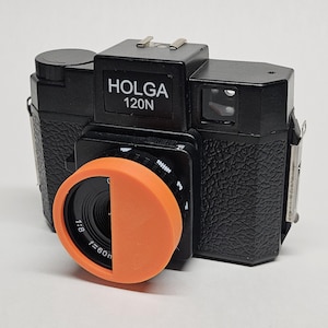 May include: A black Holga 120N camera with an orange lens cap. The camera has a textured black body and a viewfinder on the top right side. The lens cap has the text "1:8 f=60m" printed on it.