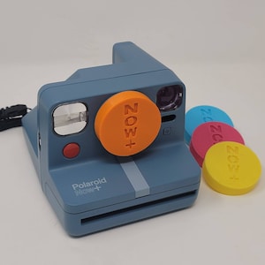 Puede incluir: Una cámara instantánea Polaroid Now+ azul con una tapa de lente naranja que dice "NOW+". Varias tapas de lente de colores están junto a la cámara. La cámara tiene un botón rojo y un flash.