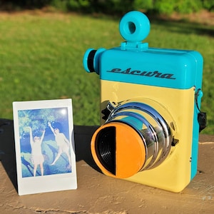 May include: A vintage-style camera in teal and yellow with the word "escura" printed on the front. The camera has a large lens with an orange accent. A small photo of two people is next to the camera.