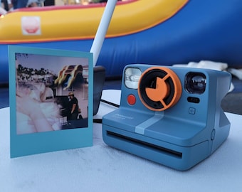 May include: A blue Polaroid camera with an orange lens and a white stripe. The camera is sitting on a white table next to a blue picture frame with a photo inside.