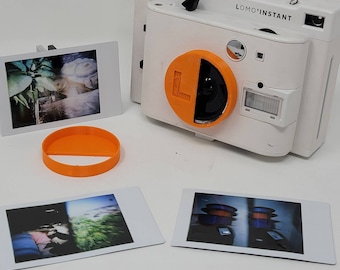 May include: A white LOMO'INSTANT camera with an orange lens cap attached. The camera is sitting on a white surface with four instant photos.