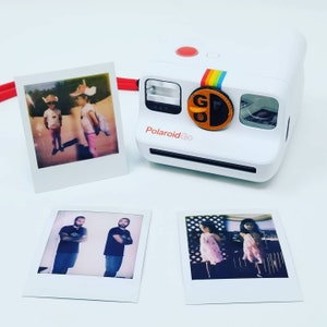 以下が含まれることがあります： 虹色のストライプと「Polaroid Go」という金色のロゴが付いた白いPolaroid Goインスタントカメラ。白い表面にPolaroidインスタント写真が4枚置かれています。写真は、子供2人、大人2人、女の子2人を写しています。