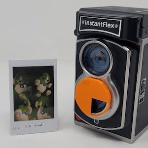 Puede incluir: Una cámara InstantFlex negra y plateada con una tapa de lente naranja. La cámara tiene un flash incorporado y un visor. Una foto Polaroid de animales de peluche verdes está en primer plano.