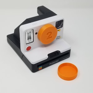 Peut inclure: Un appareil photo Polaroid OneStep 2 blanc et noir avec un capuchon d'objectif orange. Le capuchon d'objectif porte le numéro 2.