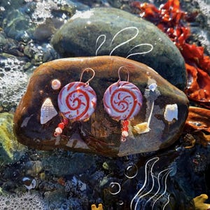 Könnte beinhalten: Ein Paar handgefertigte Ohrringe mit einem skurrilen Design. Jeder Ohrring hat eine kreisförmige, bonbonartige Form mit einem roten Spiralmuster auf hellblauem Hintergrund. Rote Perlen hängen darunter, und die Ohrringe haben kupferfarbene Haken.