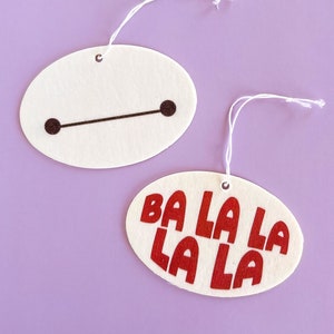 May include: Two white felt ornaments with strings for hanging. One ornament has a black line and two black circles. The other ornament has the text "BA LA LA LA LA" in red.