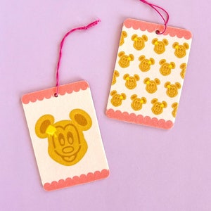 May include: Two pink and white gift tags with a Mickey Mouse theme. One tag features a single Mickey Mouse head with yellow accents, while the other tag has a repeating pattern of Mickey Mouse heads.