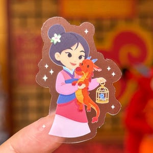 May include: A clear sticker featuring a cartoon illustration of a character in a pink and blue outfit holding a red dragon. The dragon holds a small cage. The character has dark hair and a flower. White star accents surround the image.