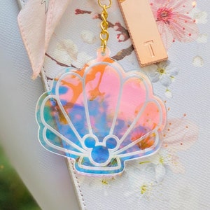 May include: A iridescent seashell-shaped keychain with a Mickey Mouse silhouette inside. The keychain is attached to a gold chain and a gold key ring.