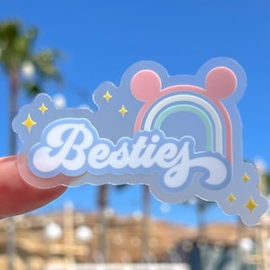 May include: A clear sticker featuring a rainbow, two pink ears, and the word "Besties" in a retro font. The sticker has a light blue background and gold glitter accents.