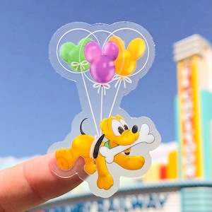 May include: A clear sticker featuring Pluto the dog holding a bone and flying with three Mickey Mouse-shaped balloons in green, purple, and yellow.