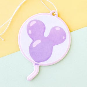 May include: A round, white ornament with a light purple Mickey Mouse balloon design. The balloon is outlined in a light pink border. A white string is attached for hanging. The background is a diagonal split of yellow and light green.