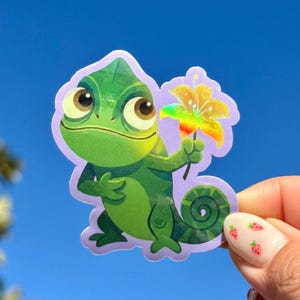 May include: A green chameleon sticker with large brown eyes and a yellow flower. The sticker has a purple border and is held against a bright blue sky. The chameleon is holding a flower with rainbow-colored petals.