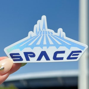 May include: A white sticker with a blue and white illustration of the Spaceship Earth ride at Epcot. The text "SPACE" is printed in blue below the illustration.