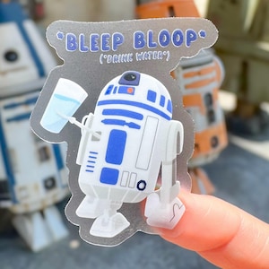 May include: A clear sticker featuring a cartoon R2-D2 droid holding a glass of water. The sticker has the text "Bleep Bloop" and "(Drink Water)" in blue. The droid is white and blue, with a small black circle on its head.