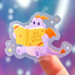 Könnte beinhalten: Ein lila Drache mit orangefarbenen Flügeln liest ein gelbes Buch mit dem Titel "Wie man eingängige Songs schreibt". Das Buch hat einen Regenbogen auf dem Cover und eine Musiknote auf jeder Seite.