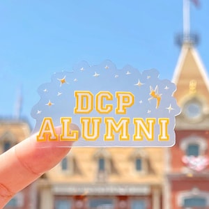 May include: A clear sticker with gold lettering that reads "DCP ALUMNI" with a small gold fairy silhouette and gold stars. The sticker is held up in front of a blurred background of a building with a red roof.