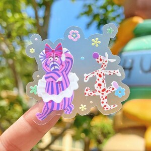 Puede incluir: Una pegatina transparente con dos personajes de dibujos animados. Uno es un gato a rayas moradas y blancas con un lazo rosa y un cuello con volantes blancos. El otro es un perro blanco con manchas rojas. Pequeñas flores amarillas, rosas y azules rodean a los personajes.