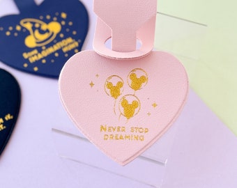May include: Pink leather heart-shaped luggage tag with a gold Mickey Mouse silhouette and the text "Never Stop Dreaming".