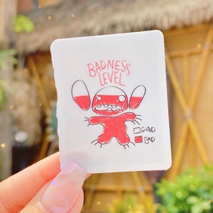 Puede incluir: Una pegatina blanca con un personaje de dibujos animados rojo y el texto "Badness Level". El personaje tiene ojos grandes y una boca ancha. Hay dos casillas debajo del personaje, una etiquetada como "Good" y la otra como "Bad".