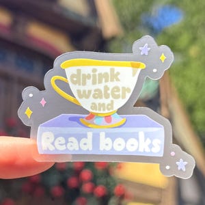 以下が含まれることがあります： 「drink water and Read books」の文字が入った、ティーカップのデザインのクリアステッカー。 ティーカップは黄色と白で、紫色の台座が付いています。 ステッカーは星の付いた気まぐれなデザインです。