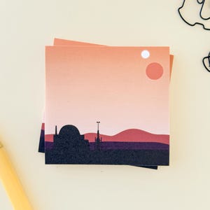 May include: A stack of square notepads featuring a sunset landscape with a silhouette of a city. The color palette includes shades of orange, pink, purple, and black. A pen and paperclips are also visible.