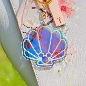 May include: A iridescent seashell keychain with a Mickey Mouse silhouette inside. The keychain is attached to a gold chain and a gold key ring.