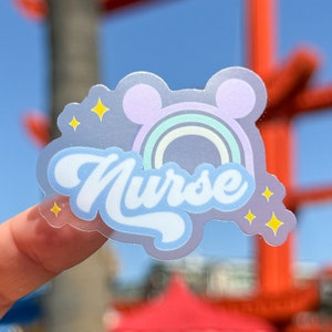 May include: A clear sticker with a pastel rainbow, Mickey Mouse ears, and the word "Nurse" in a whimsical font. The sticker has a light blue border and is decorated with yellow star accents. The background is a blurred outdoor scene.