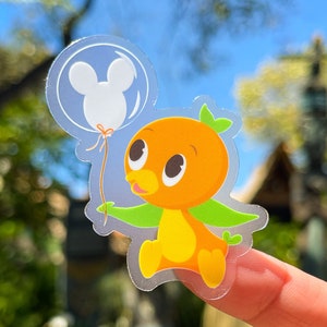 May include: A clear sticker of a cartoon orange bird holding a balloon with a Mickey Mouse silhouette. The bird is wearing a green leaf on its head and has a yellow body.