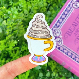 Puede incluir: Una pegatina blanca con una taza de crema batida de estilo caricatura. La taza tiene un asa y un borde amarillos, con una base morada y azul. La crema batida es de color marrón claro con chispas blancas. La pegatina está sujeta por un dedo.