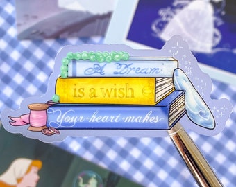 Puede incluir: Una pegatina con una pila de libros que dice "A Dream is a wish your heart makes". La pegatina está sobre un fondo a cuadros azul y blanco.