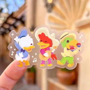May include: A clear sticker featuring three cartoon characters playing music. The characters are a duck wearing a blue bow tie, a rooster wearing a sombrero and playing a guitar, and a green bird wearing a straw hat and playing maracas.