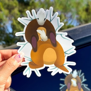 May include: A sticker featuring a cartoon animal hugging another animal, set against a white snowflake design. The animals are brown and tan with white accents. A second sticker is visible on a dark blue surface.