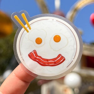May include: A clear sticker featuring a cartoon illustration of a plate of rice, two fried eggs, and bacon. The eggs and bacon are arranged to form a smiley face. Two yellow chopsticks are sticking out of the top of the plate.