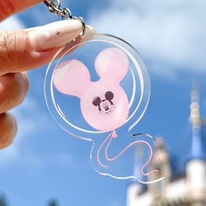 Op de afbeelding: Een transparante acryl sleutelhanger met een roze Mickey Mouse ballon met een zwarte omtrek. De ballon is gevormd als Mickey Mouse's hoofd en heeft een zwart gezicht met witte ogen en een zwarte neus.