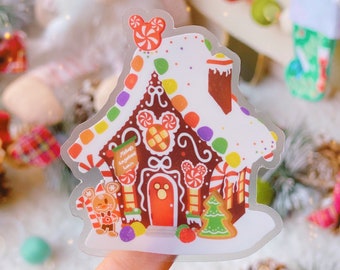 May include: A clear sticker of a gingerbread house with white frosting and colorful candy decorations. The house has a red door, a chimney, and a sign that reads "Mickey's Gingerbread House". A gingerbread man and a Christmas tree are also featured.