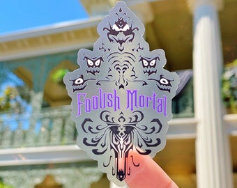 Könnte beinhalten: Ein transparenter, schimmernder Aufkleber mit einem schwarz-weißen Design des Haunted Mansion-Logos. Das Logo zeigt einen Geist mit einer Krone und den Text "Foolish Mortal".