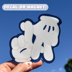 May include: White cartoon-style hands with a blue outline, forming the shape of the letter "L" and the number "2". The text "DECAL OR MAGNET!" is above the hands. The background is a clear blue sky.