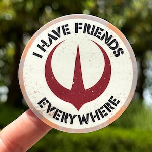 May include: A round sticker with the text "I HAVE FRIENDS EVERYWHERE" in black, surrounding a maroon symbol. The sticker has a distressed, vintage look with an off-white background and an orange border. The sticker is held by a finger.