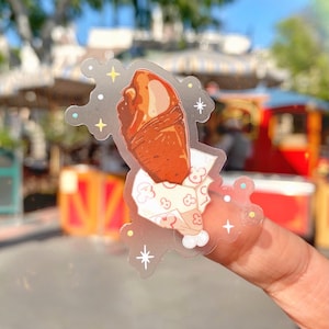 May include: A clear sticker featuring a cartoon illustration of a turkey leg. The turkey leg is brown with a white and pink paper wrap. The sticker has a decorative border with stars and sparkles.