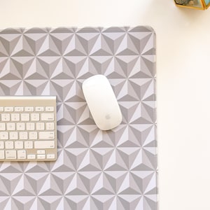 May include: A computer mouse and keyboard rest on a geometric patterned mousepad. The mousepad features a repeating 3D triangle design in shades of gray. The keyboard is silver and the mouse is white.
