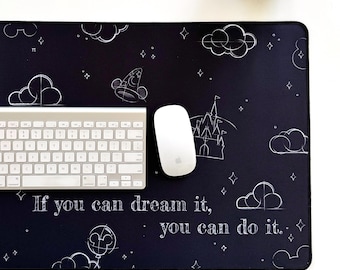 May include: A black rectangular desk mat with white sketched illustrations of clouds, a castle, a wizard hat, and a Mickey Mouse balloon. The mat has the quote, "If you can dream it, you can do it." A white keyboard and mouse are on the mat.