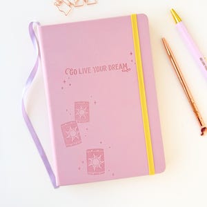 Puede incluir: Un diario lila con el texto "Go Live Your Dream" e ilustraciones de linternas. Tiene una banda elástica amarilla y una cinta lila. También hay un bolígrafo de oro rosa, un bolígrafo lila y un clip con forma de castillo.