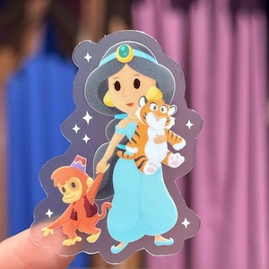 May include: A clear sticker featuring a cartoon illustration of Princess Jasmine in a blue dress, holding a tiger and a monkey. The sticker has a blue headband with a gold jewel and is surrounded by white stars.
