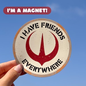 Può includere: Magnete rotondo con un design invecchiato. Il magnete presenta un emblema rosso e bianco con le parole "I HAVE FRIENDS EVERYWHERE" in testo nero. Un banner in alto recita "I'M A MAGNET!" in testo bianco su sfondo marrone.