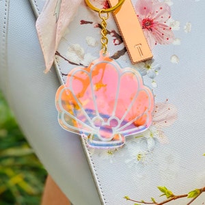May include: A iridescent pink seashell keychain with a Mickey Mouse silhouette inside. The keychain is attached to a gold chain and a gold key ring.
