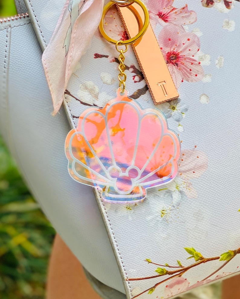 May include: A iridescent pink seashell keychain with a Mickey Mouse silhouette inside. The keychain is attached to a gold chain and a gold key ring.